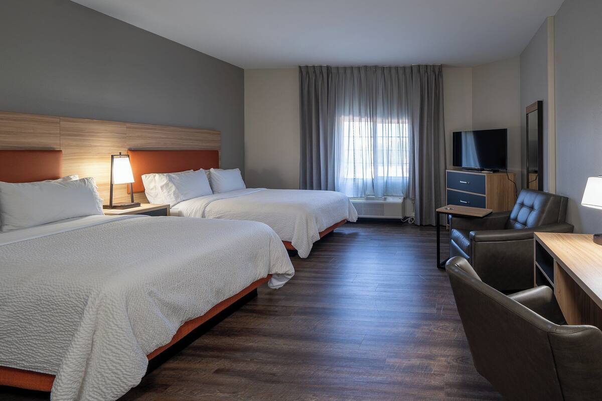 A hotel room with two beds, a desk, a chair, a TV, and an air conditioning unit.