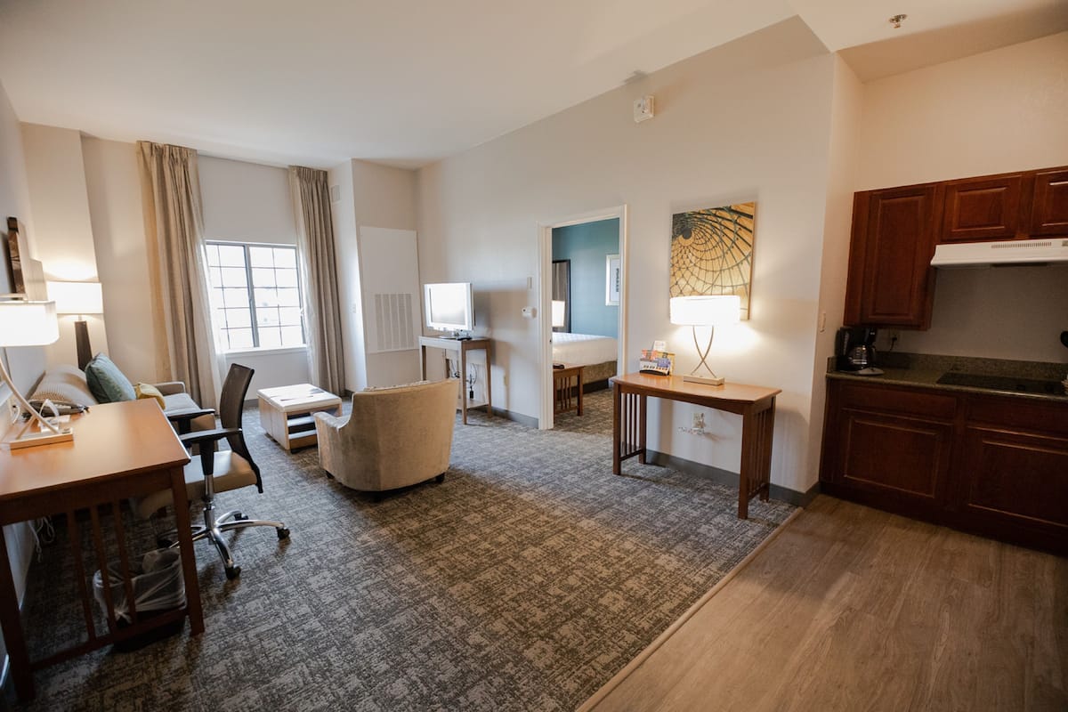 A hotel room with a living area, a dining table, a sofa, a small kitchen, and a window with curtains.