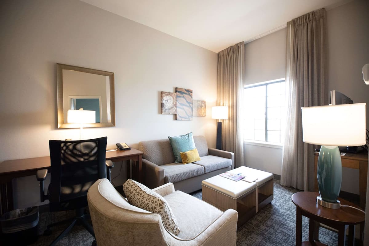 Une chambre d’hôtel équipée d’un canapé, d’un fauteuil, d’un bureau et d’une lampe. On y trouve une fenêtre avec des rideaux, un miroir et des œuvres d’art mural.