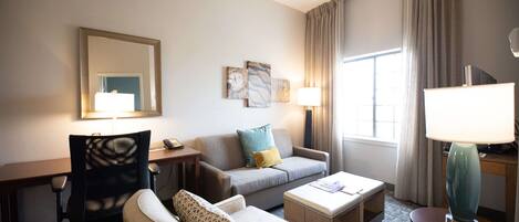 A hotel room with a sofa, armchair, desk, and lamp. There is a window with curtains, a mirror, and wall art.