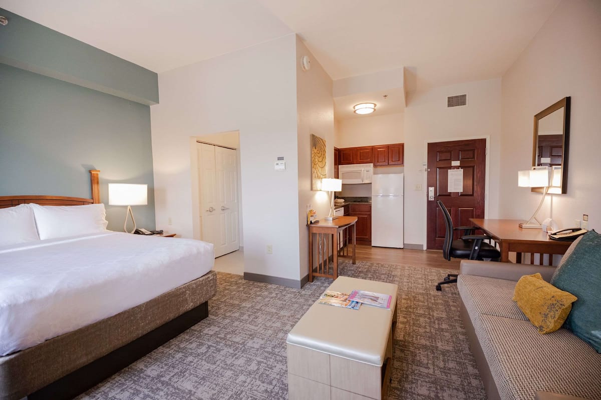 Une chambre d’hôtel avec un lit, une table de chevet, une lampe et une petite table sur laquelle se trouve un magazine. Il y a également une kitchenette équipée d’un four à micro-ondes, d’une cuisinière et d’un réfrigérateur. On peut aussi apercevoir un canapé et un fauteuil.