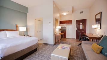 Habitación de hotel con cama, mesita de noche, lámpara y una pequeña mesa con una revista. Se observa una kitchenette con microondas, estufa y refrigerador. También se puede ver un sofá y una silla.