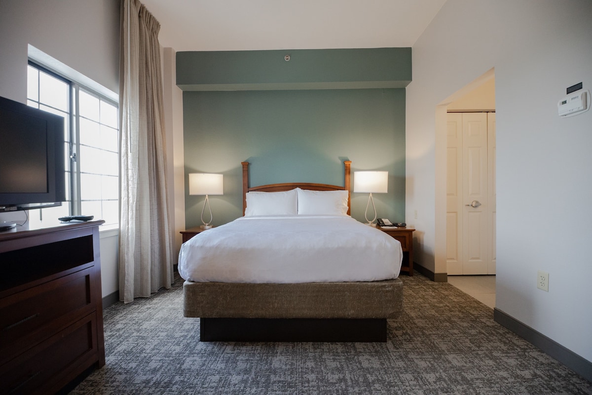 Una habitación de hotel con una cama grande, dos lámparas de noche, un televisor sobre un aparador y una ventana con cortinas.
