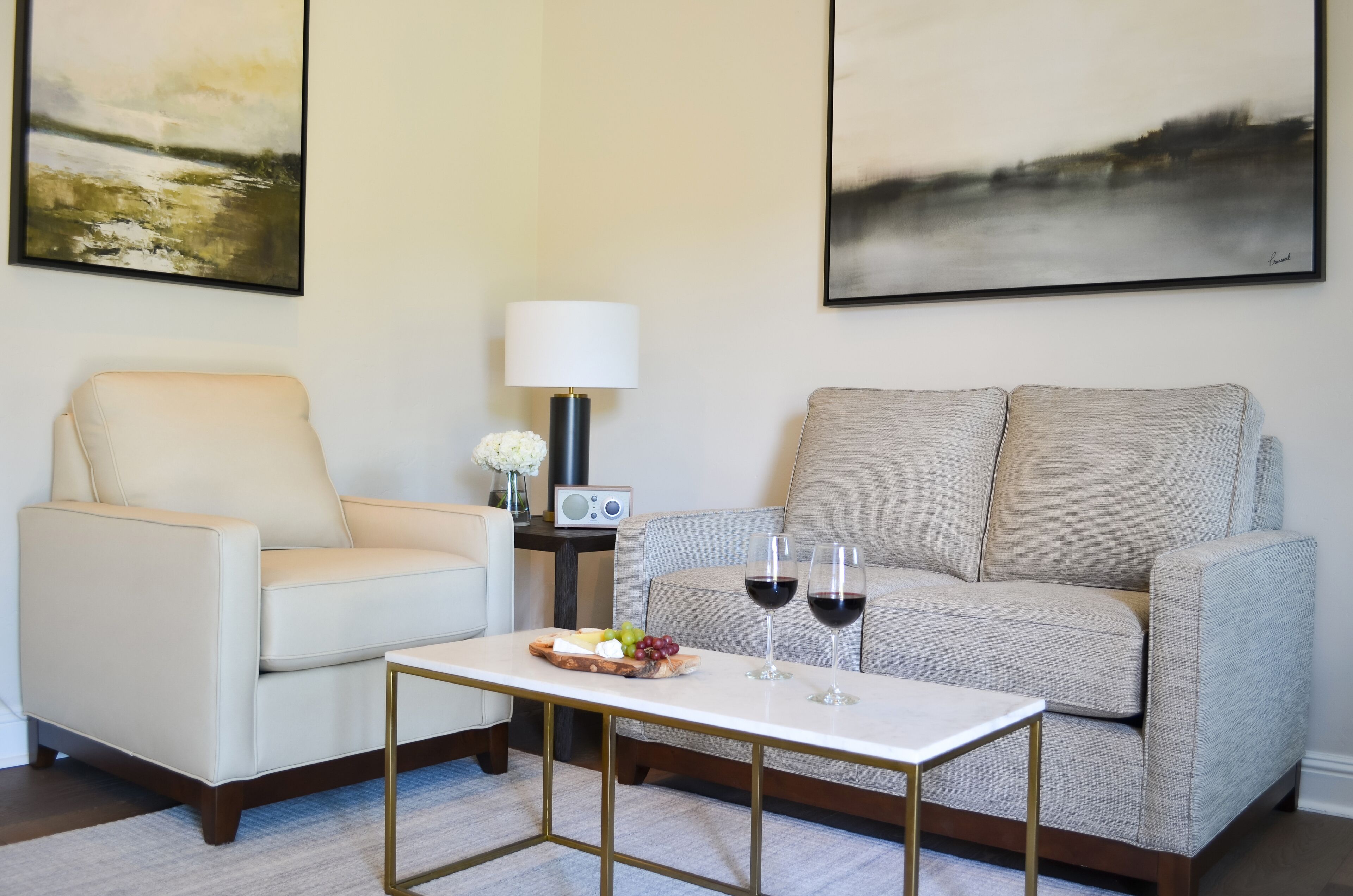 Ein Wohnzimmer mit einem beigen Sessel, einem grauen Sofa, einem kleinen Tisch mit einer Obstplatte und zwei Weingläsern sowie einer Lampe.