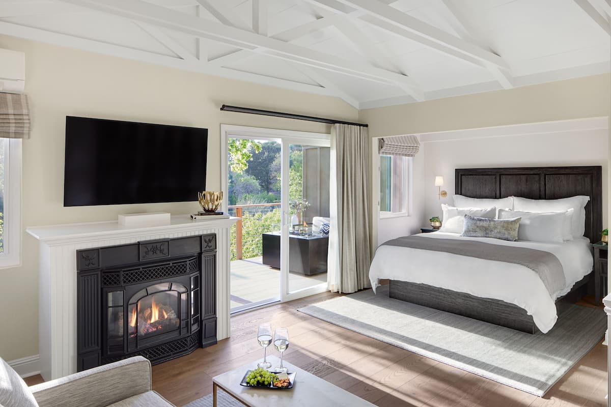A spacious bedroom with a fireplace, a large bed, a television, and a balcony with a view of greenery.