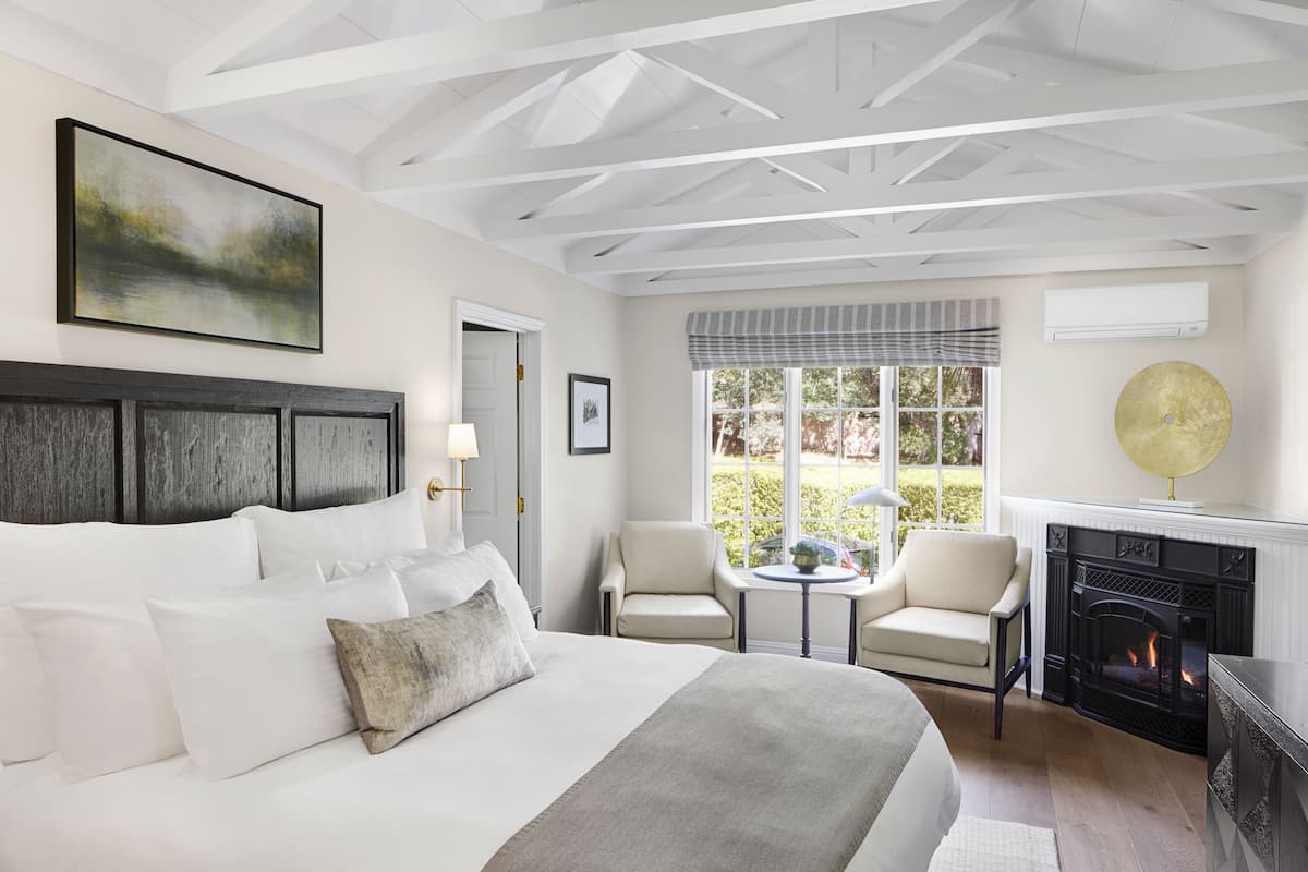 A well-lit bedroom with a large bed, a fireplace, a dining table with chairs, and a view of the outdoors.
