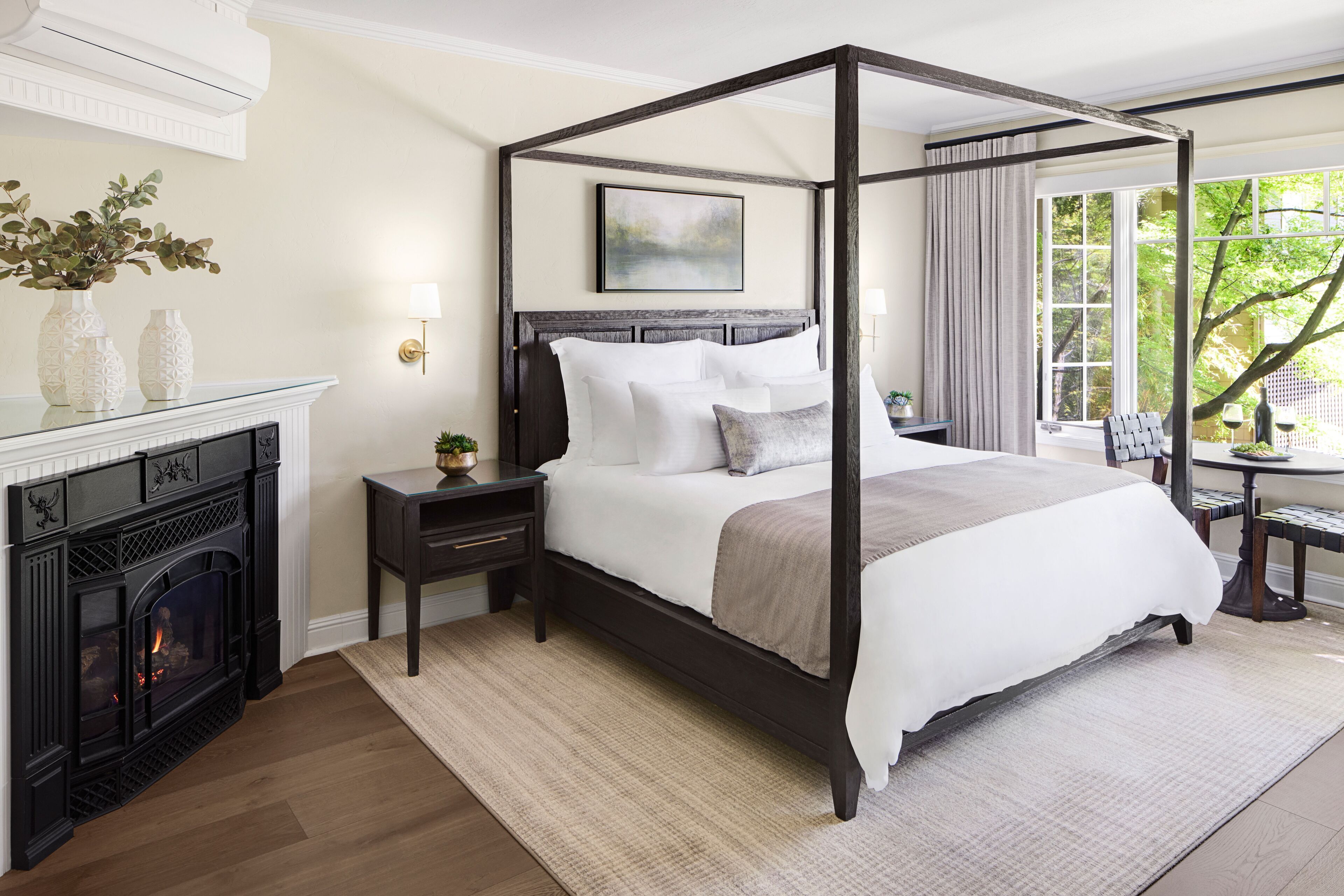 A four-poster bed with a dark wooden frame, white bedding, and a beige throw blanket. There is a fireplace with a mantel and decorative vases on the left. A small table with a lamp and a chair is visible to the right of the bed.