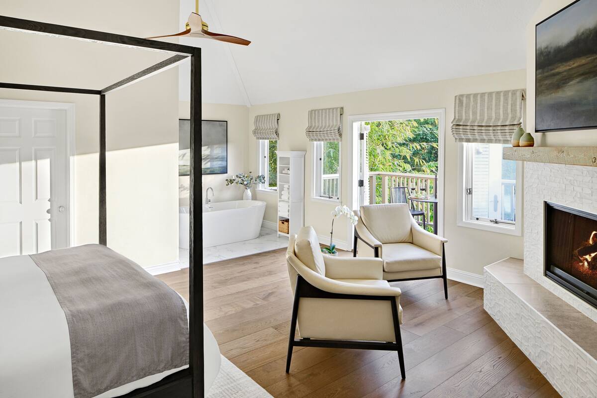 A spacious bedroom with a large bed, a sitting area with chairs, a fireplace, and a balcony with a view of greenery.