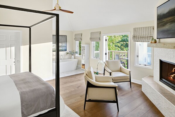 A spacious bedroom with a large bed, a sitting area with chairs, a fireplace, and a balcony with a view of greenery.