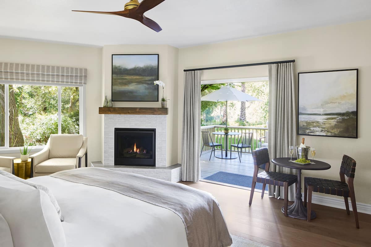 A bedroom with a fireplace, a balcony with outdoor seating, and a view of greenery.