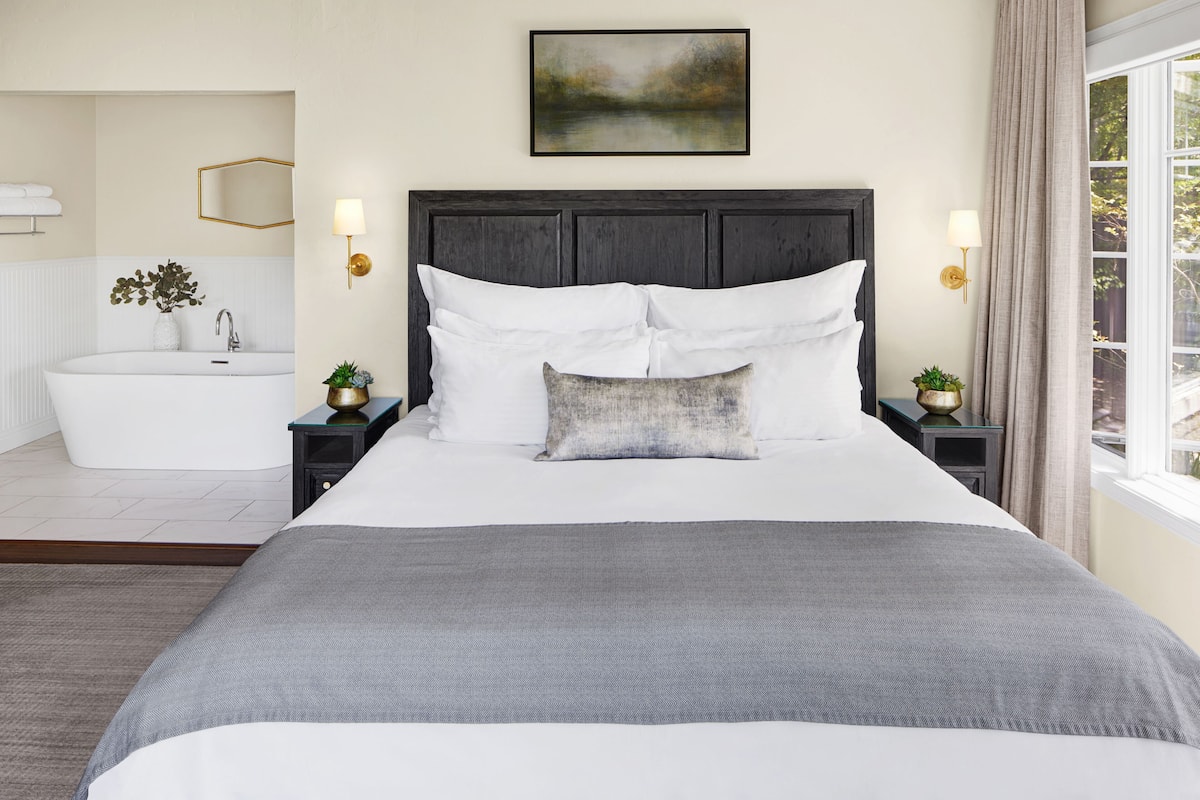 A bedroom with a large bed, a nightstand with a lamp, a wall-mounted painting, and a view of a bathroom with a bathtub and a sink.