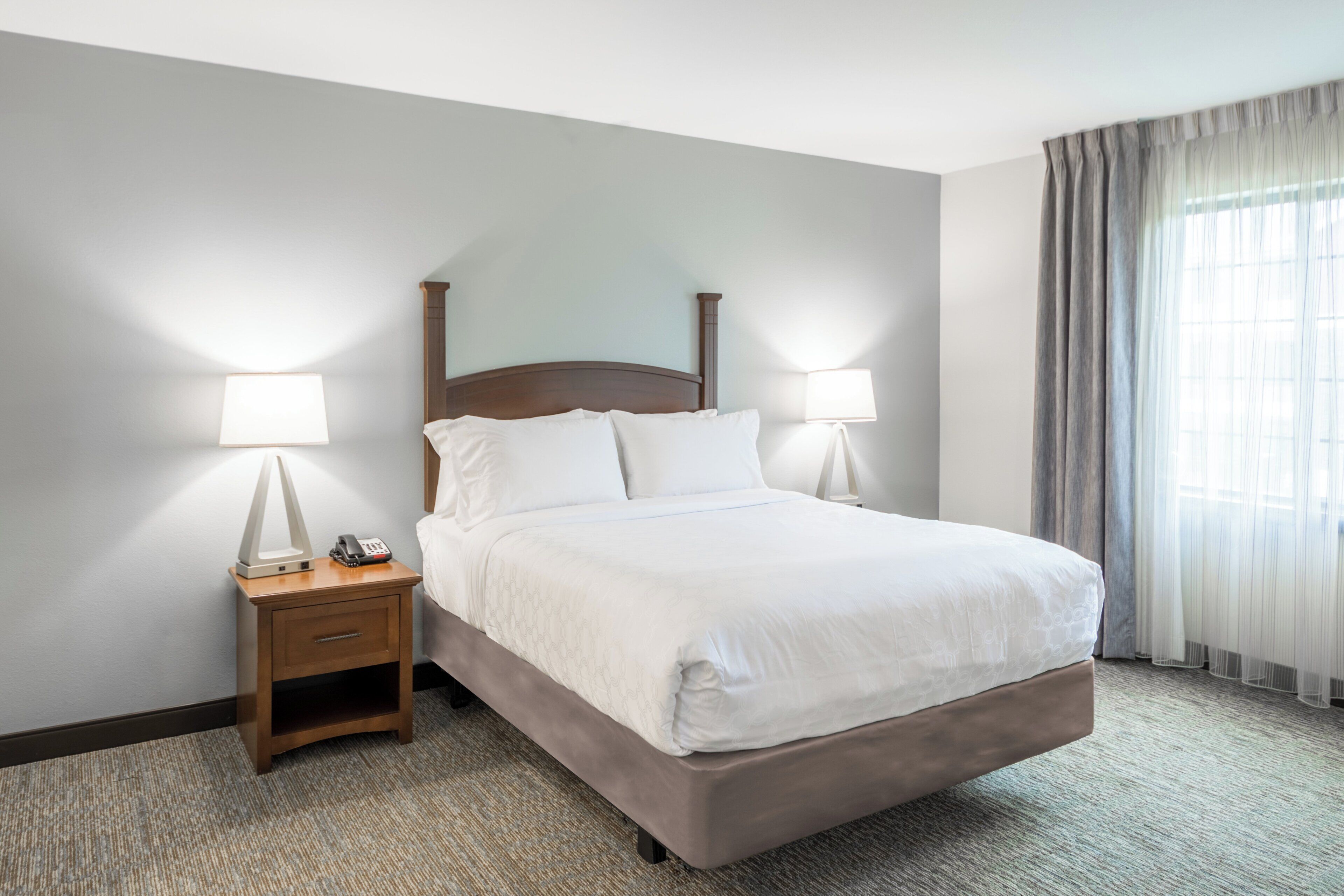 Habitación de hotel con cama, dos lámparas de noche, mesita de noche con teléfono y ventana con cortinas.