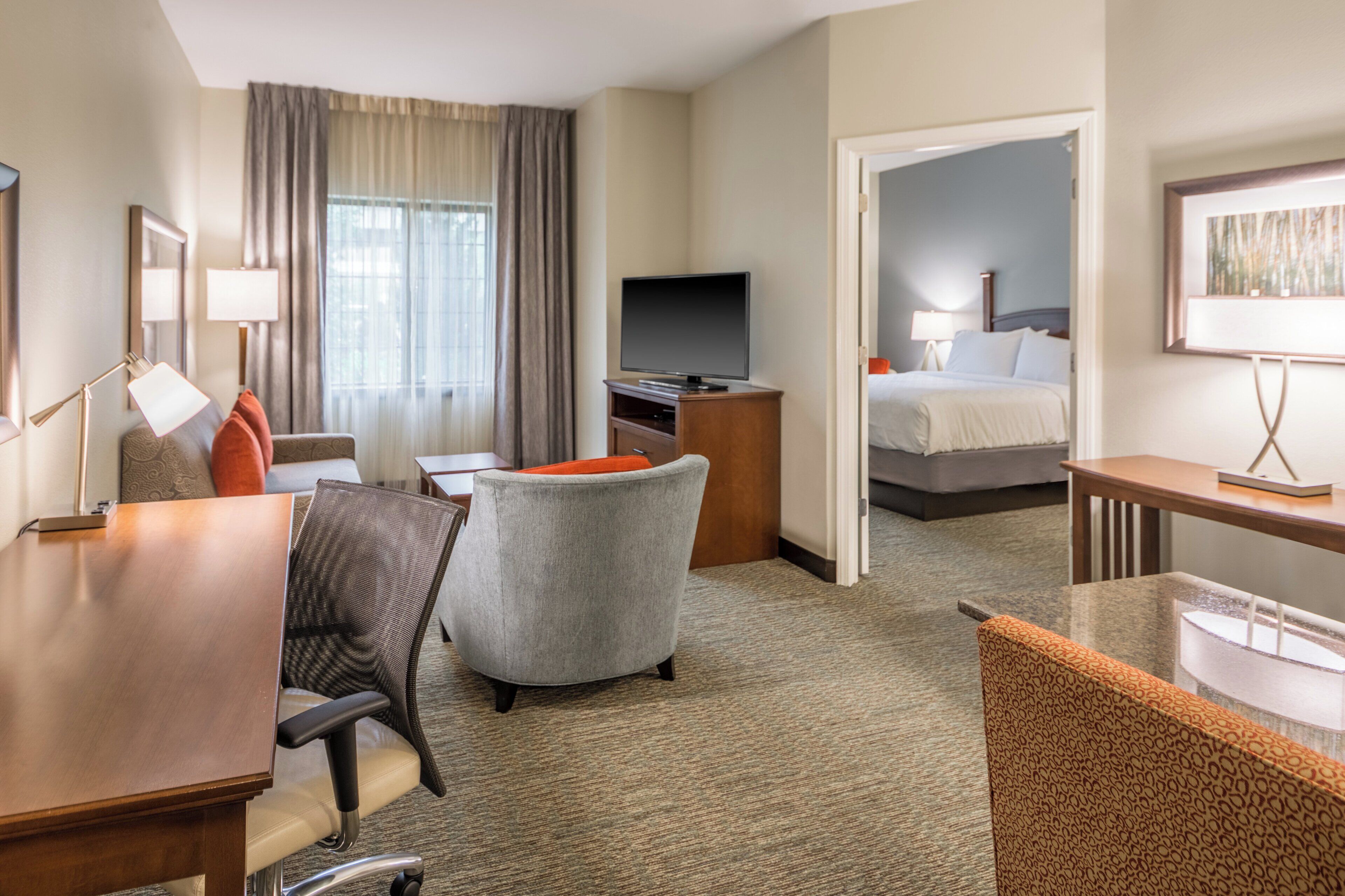 A hotel room with a desk, chair, television, bed, and a painting on the wall.