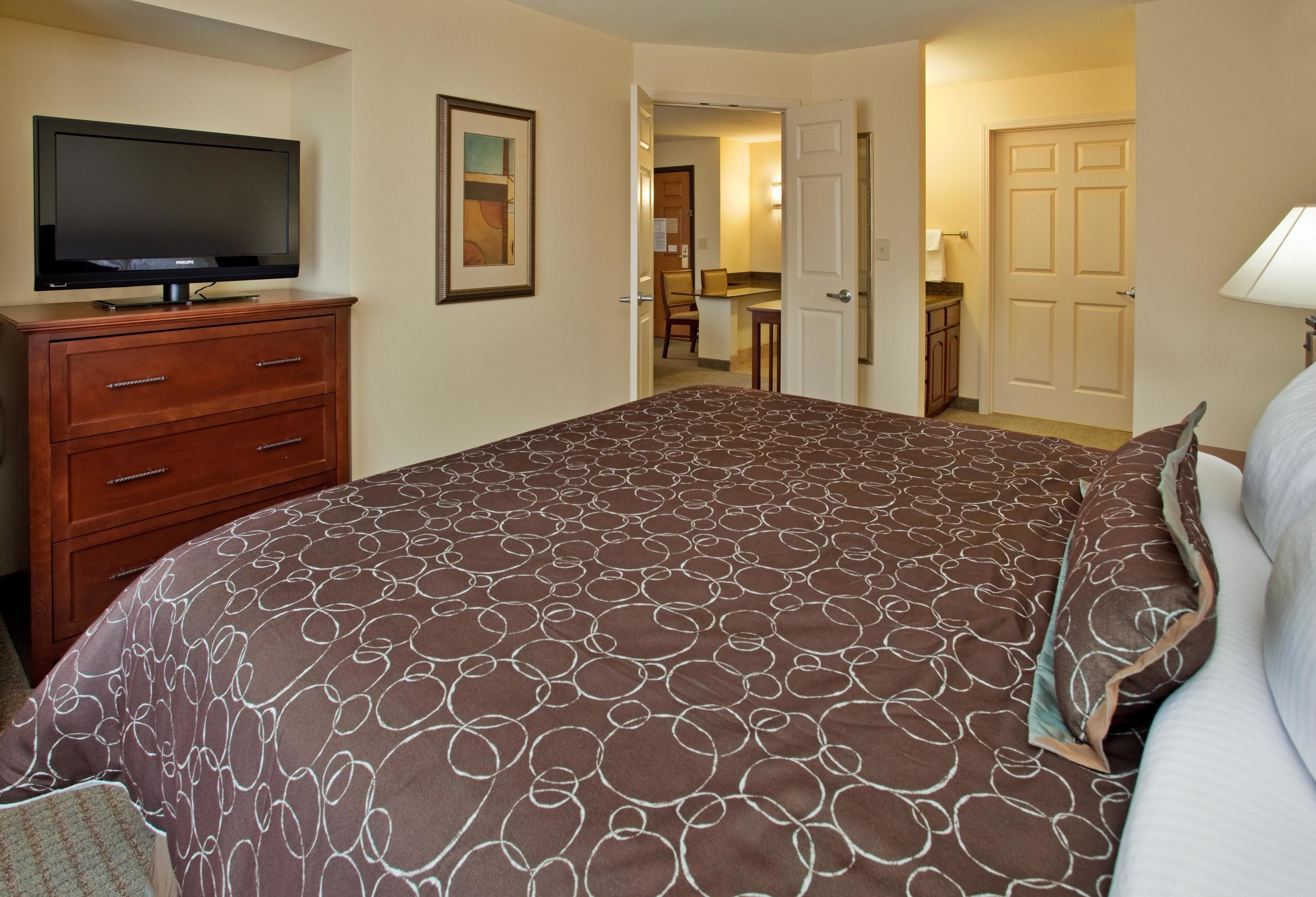 A hotel room with a large bed, a TV on a wooden dresser, a painting on the wall, and a door leading to a bathroom.