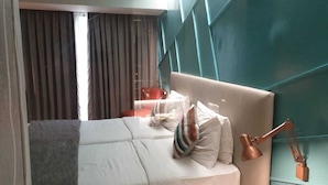 A modern bedroom with a bed, pillows, a desk lamp, and a window with curtains. - Yogi Metropolitan (Navi Mumbai)