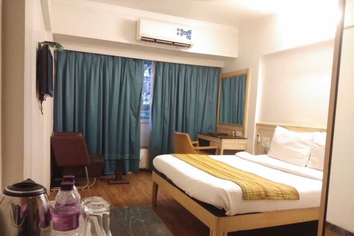 Habitación de hotel con cama, escritorio, silla, ventana con cortinas, aire acondicionado y una mesita con hervidor y botella.