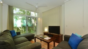 A living room with a sofa, a coffee table, a TV, and a balcony with a view of trees. - Seascape Holidays - Tropic Sands (Port Douglas)
