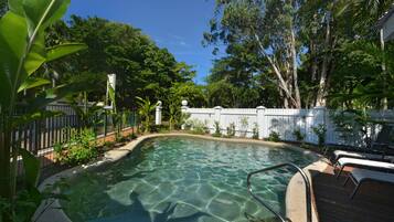 A clear swimming pool surrounded by lush greenery and a wooden deck with lounge chairs.