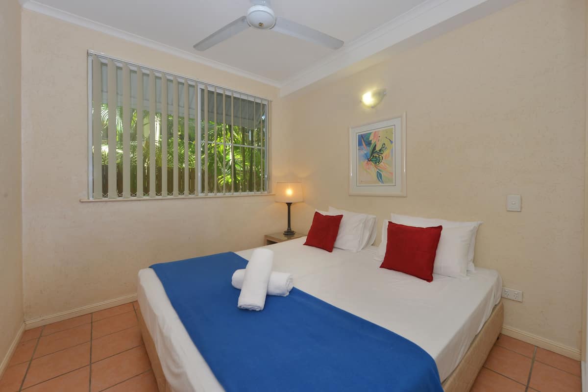 Une chambre d’hôtel avec un lit, un ventilateur de plafond, une fenêtre avec des barreaux, une table de chevet avec une lampe et un tableau au mur.
