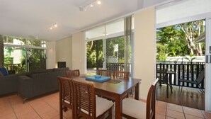 A dining area with a wooden table and chairs, a grey sofa, and a large sliding glass door leading to a balcony with a view of greenery. - Seascape Holidays - Tropic Sands (Port Douglas)
