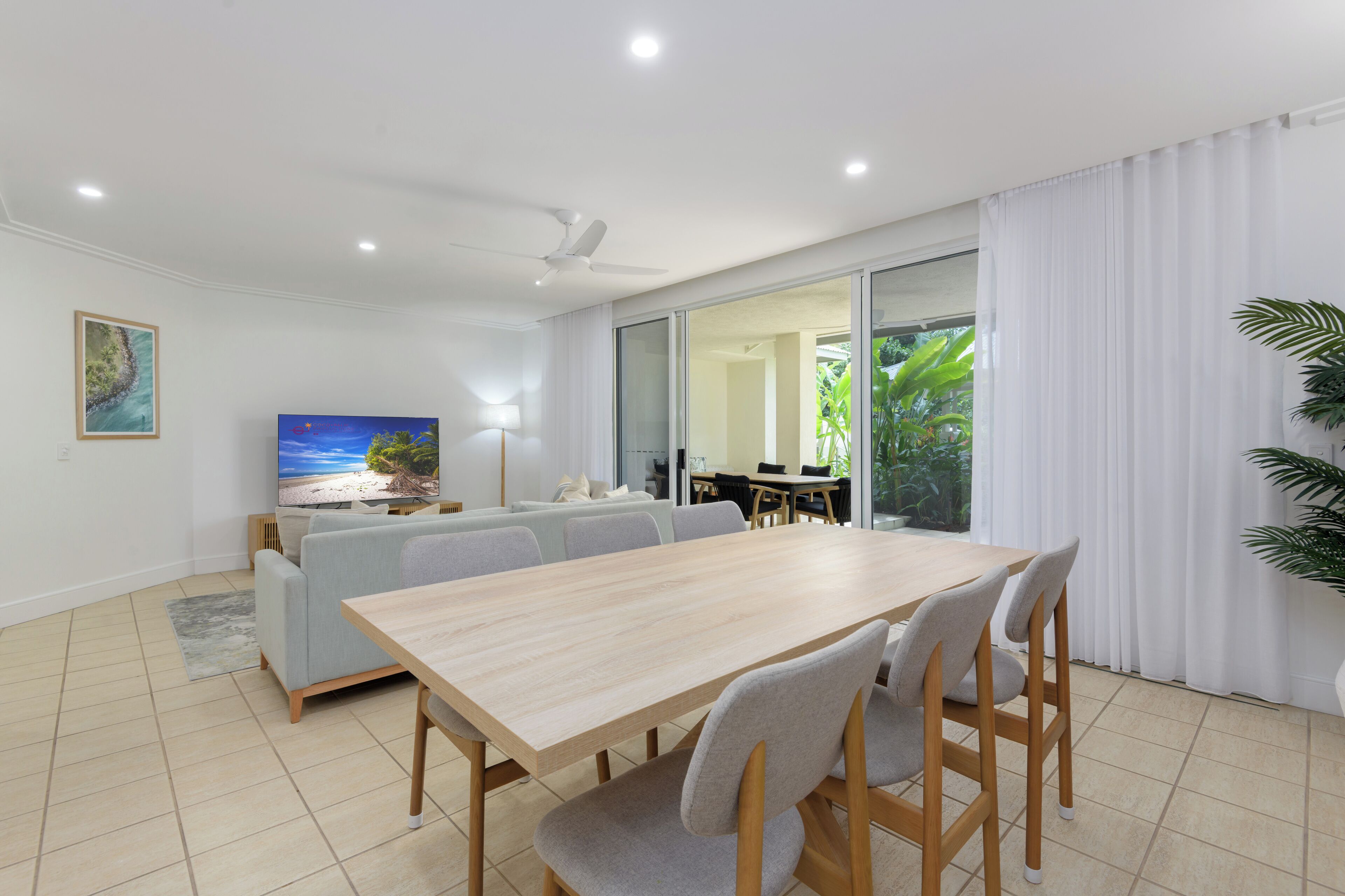 A modern living room with a wooden dining table, grey sofas, and a flat-screen TV.