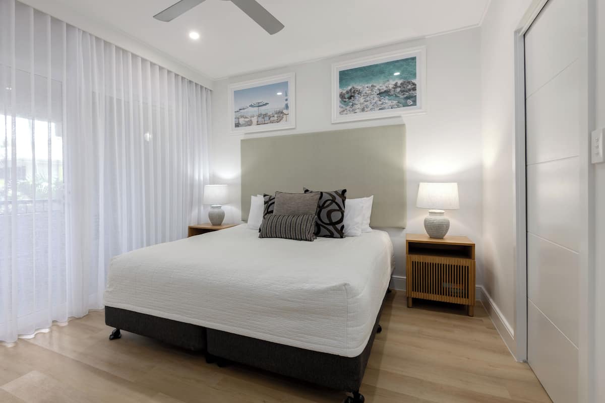 Una habitación de hotel moderna con una cama grande, dos lámparas de noche y vistas al exterior a través de cortinas translúcidas.