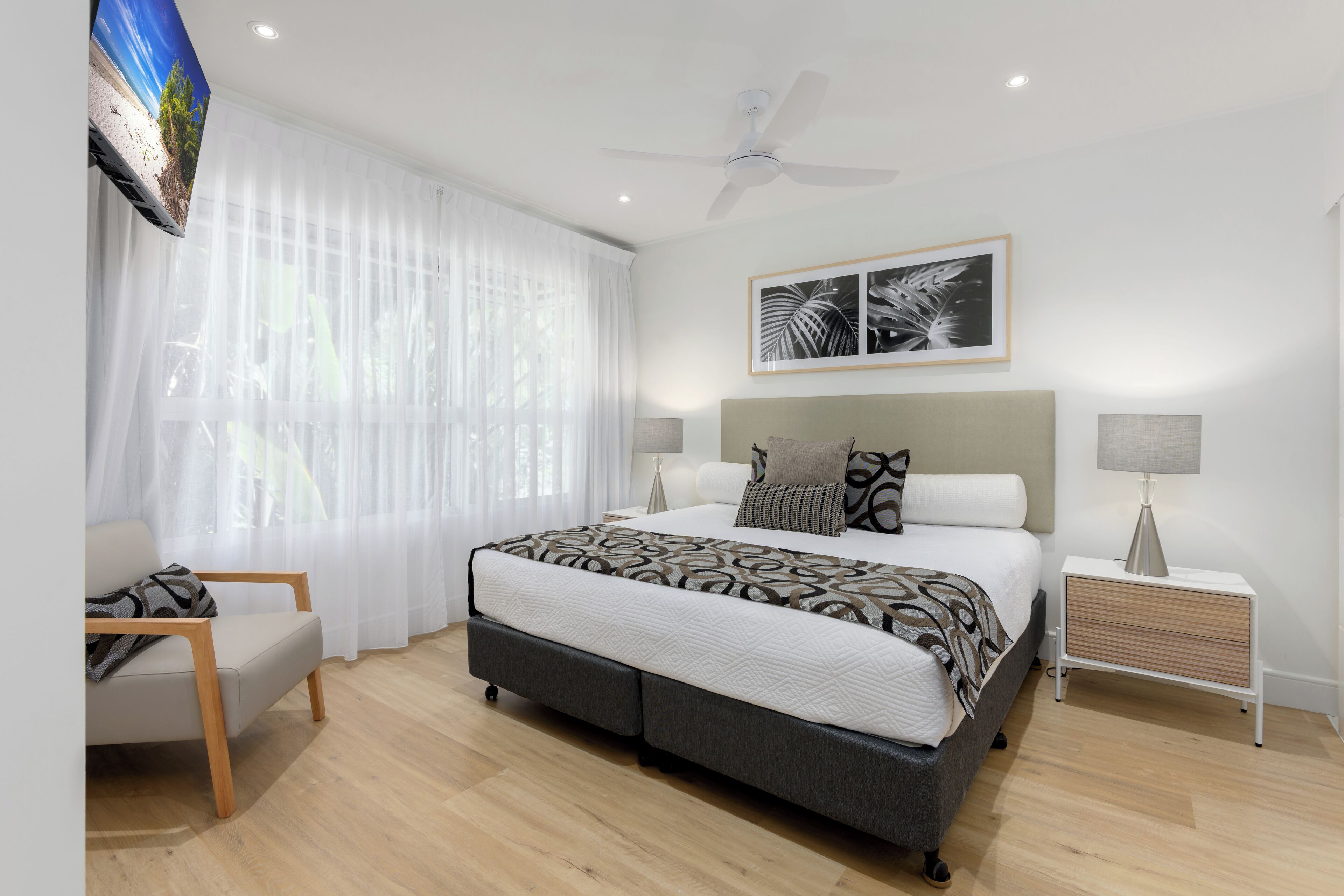 Quarto moderno com cama, cadeira, mesa de cabeceira e ventoinha no teto.