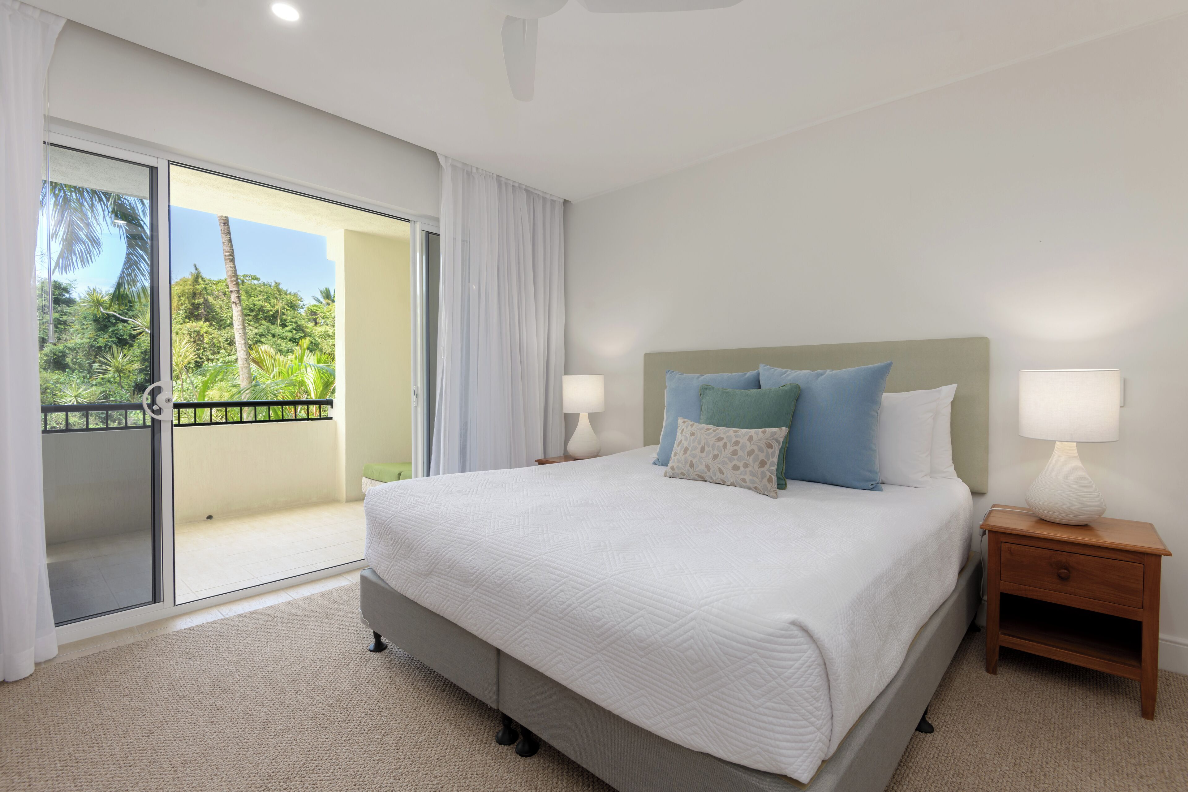 A bedroom with a large bed, a nightstand, a sliding glass door to a balcony, and a view of greenery.
