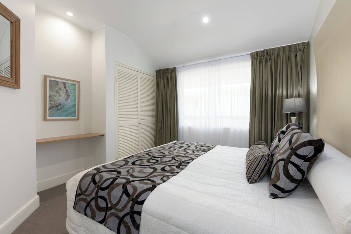 Un dormitorio ordenado con una cama grande, una lámpara de noche y una ventana con cortinas.