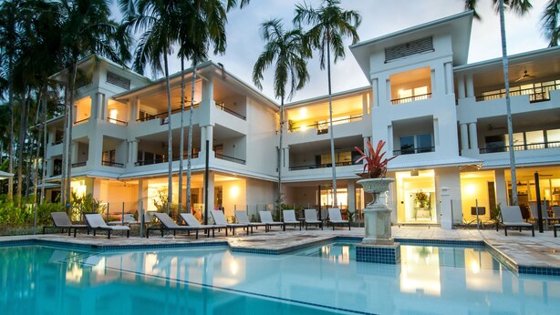 Un hotel de varios pisos con piscina, sillas de descanso y palmeras.