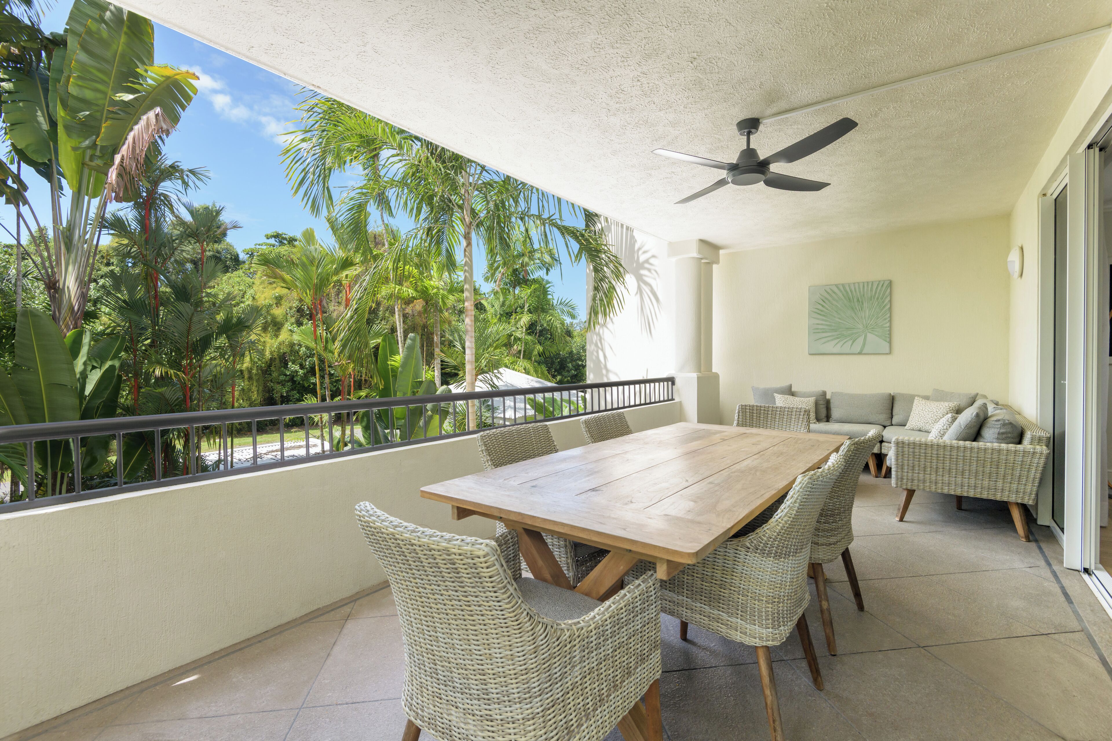 Un balcon avec une table en bois et des chaises en osier, donnant sur un jardin tropical agrémenté de palmiers.
