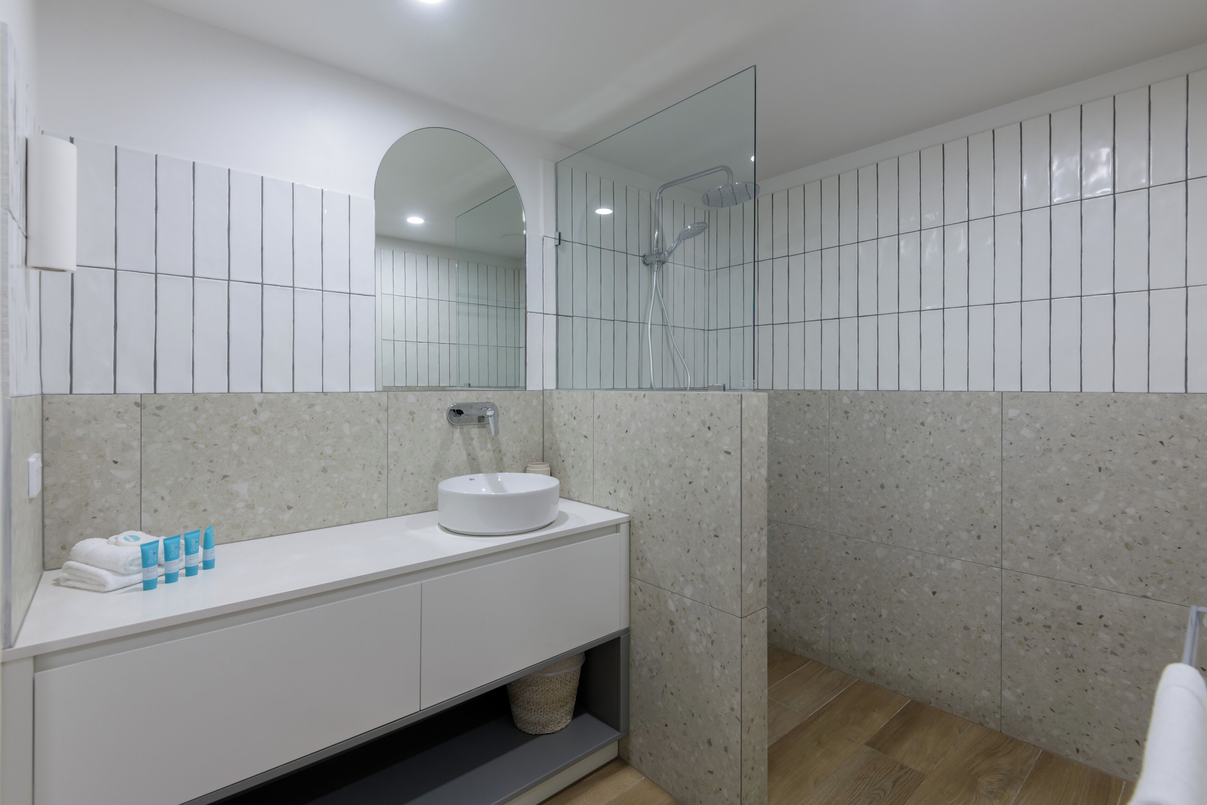 Un baño moderno con un tocador blanco, un lavabo redondo, una ducha con mampara de vidrio y un espejo grande.