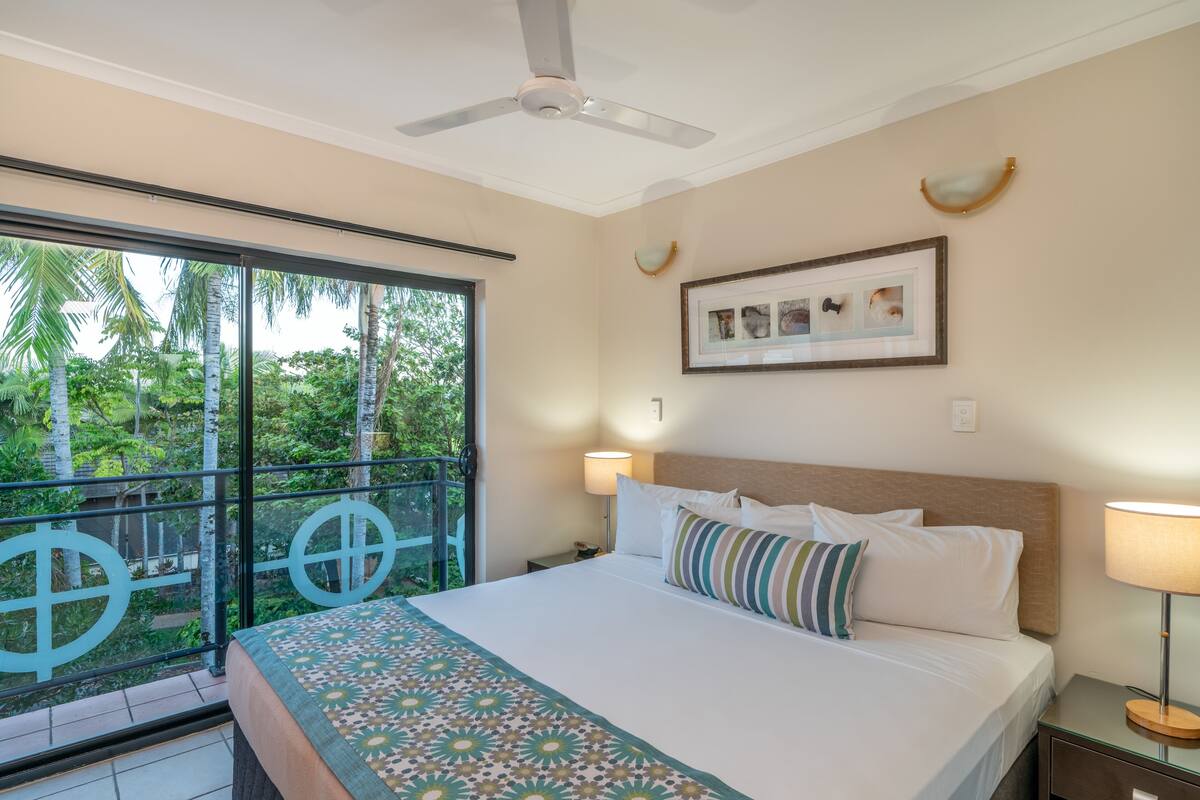 Une chambre d’hôtel avec un grand lit, un ventilateur de plafond, un tableau encadré au mur et une vue sur les palmiers par la fenêtre.