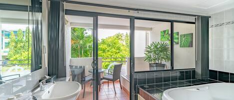 A modern bathroom with a large bathtub, a sink, and a glass-enclosed balcony with chairs and a potted plant.