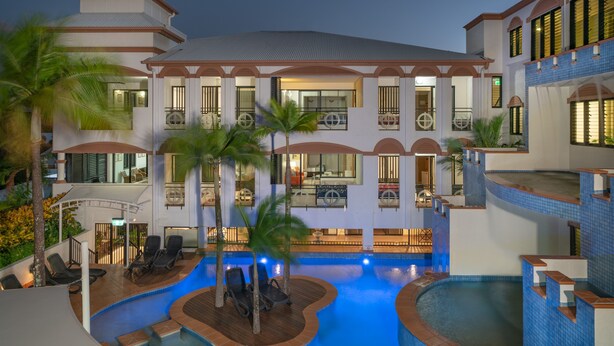 Un hotel de varios pisos con piscina, mobiliario exterior y una zona cubierta con palmeras.
