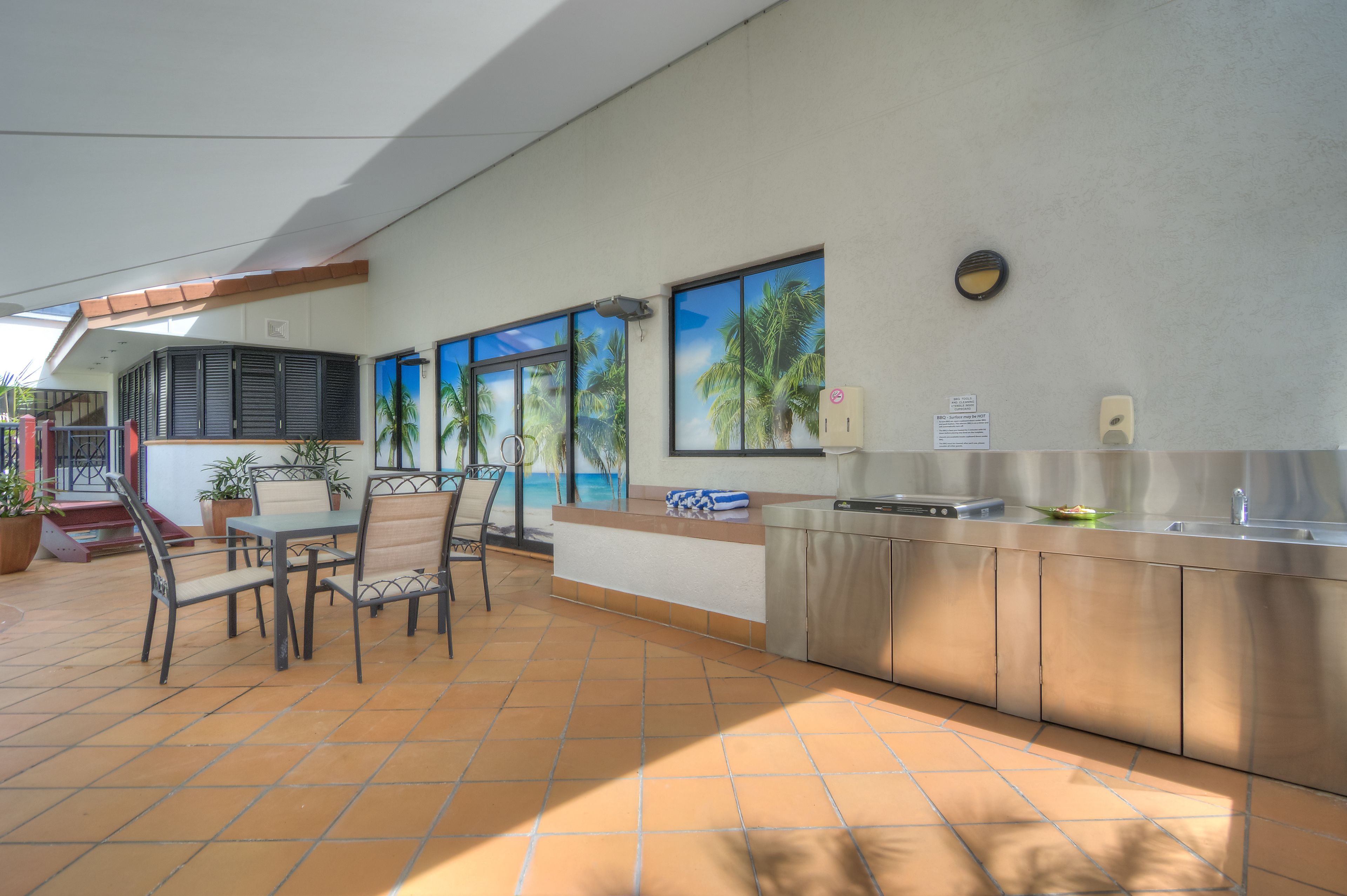 A modern kitchen with a stainless steel countertop, a dining area with chairs, and a view of palm trees through large windows.