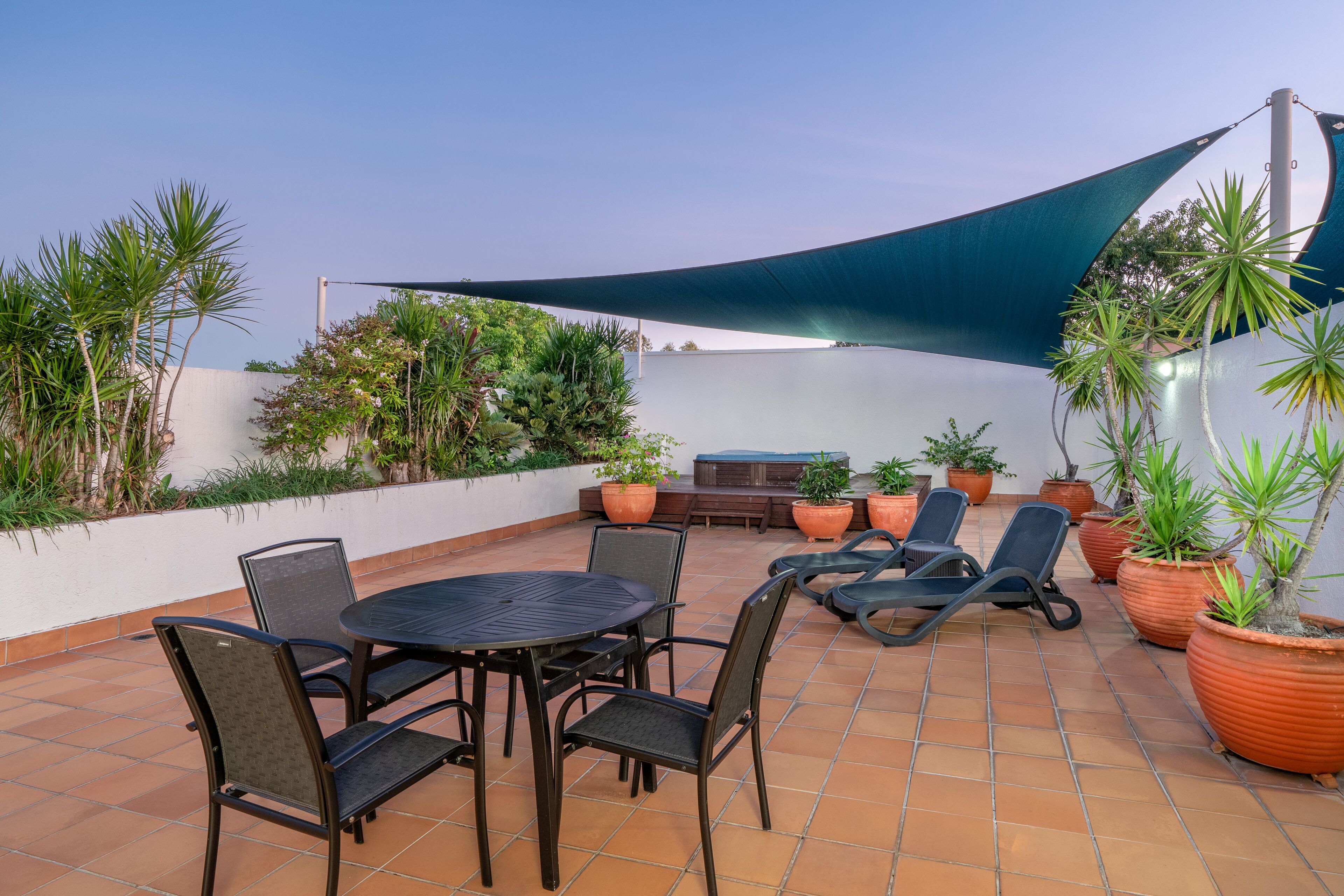 Un patio al aire libre con una mesa redonda y sillas, rodeado de plantas en macetas y una gran vela de sombra.