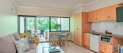 Una cocina moderna con armarios de madera, un refrigerador blanco y un microondas. Hay un comedor con una mesa de vidrio y sillas, además de un sofá con cojines estampados.