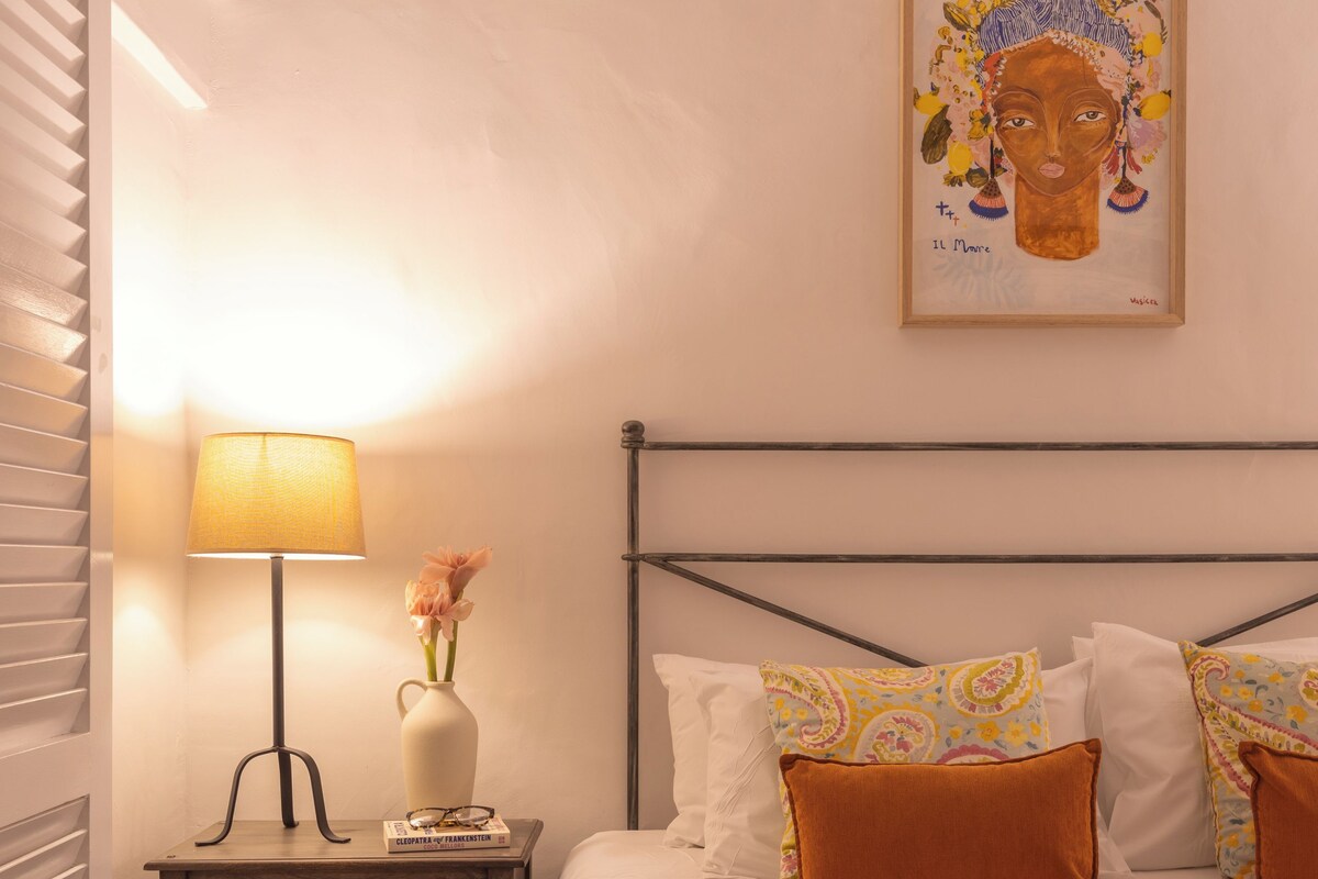 A neatly made bed with patterned bedding, a bedside lamp, a vase with flowers, and a framed artwork on the wall.