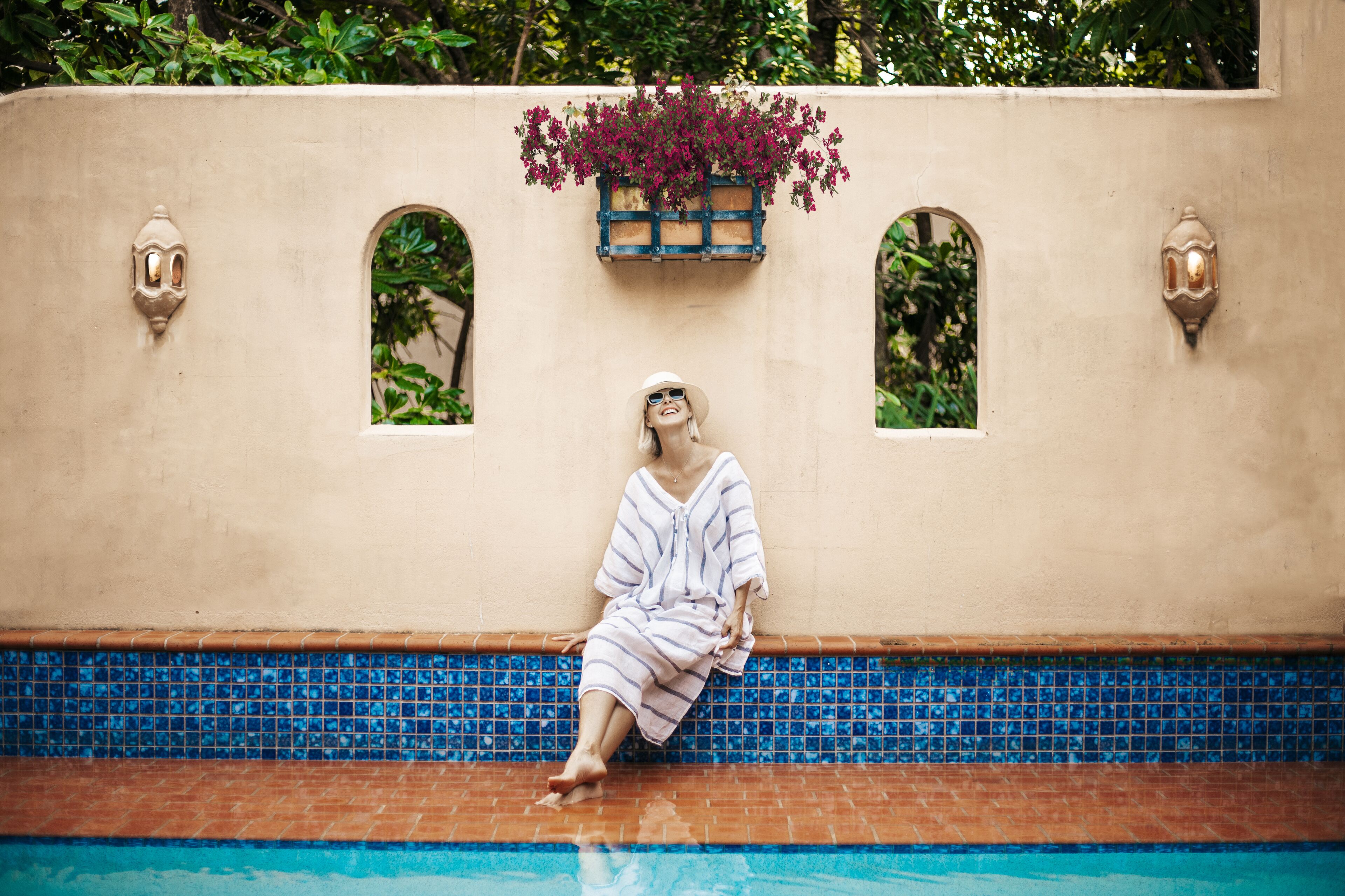 A person sitting by a pool with a tiled edge, a wall with two arched windows, and a planter with flowers above the windows.