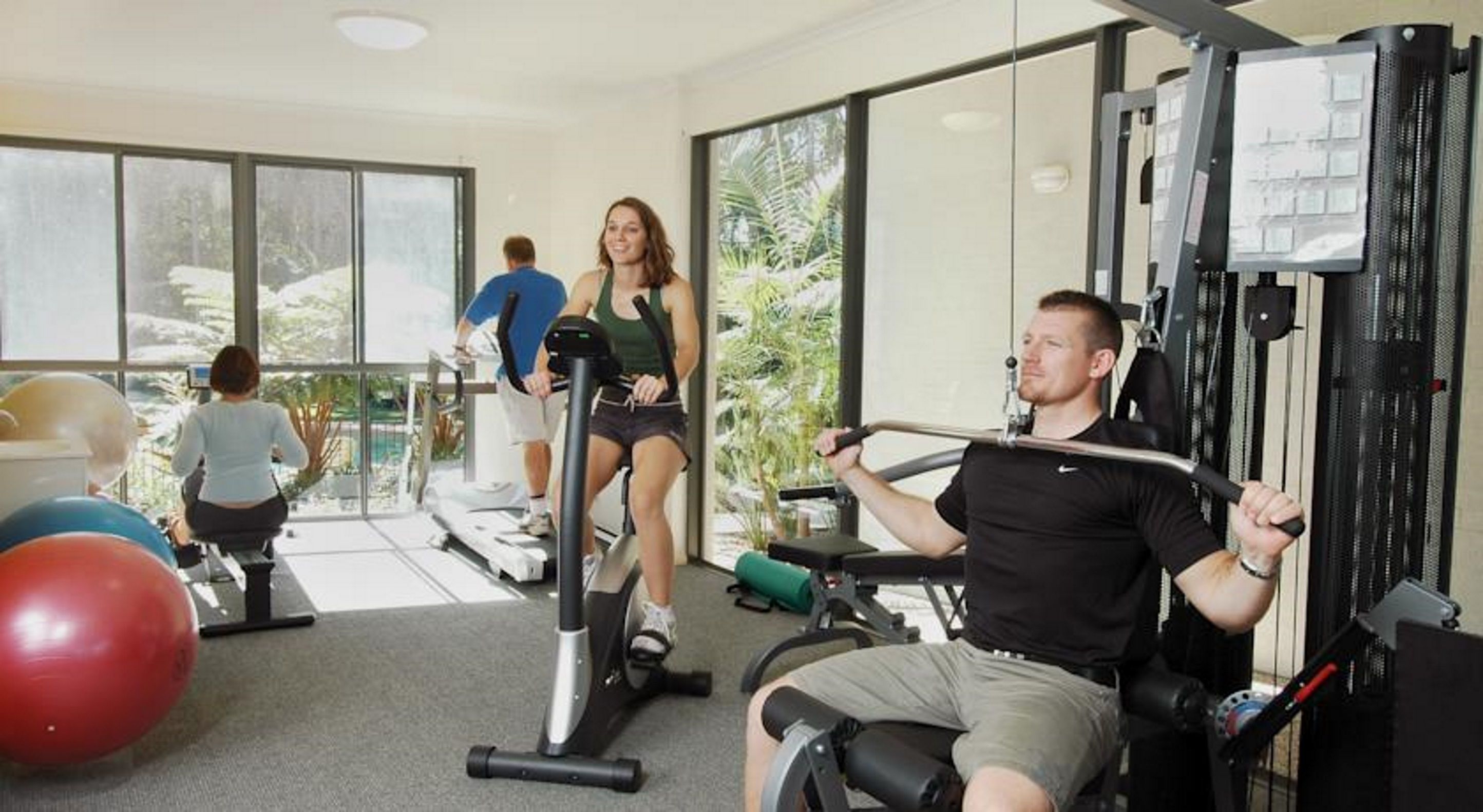 Et fitnesscenter med træningsudstyr, store vinduer og udsigt til grøn vegetation udenfor.