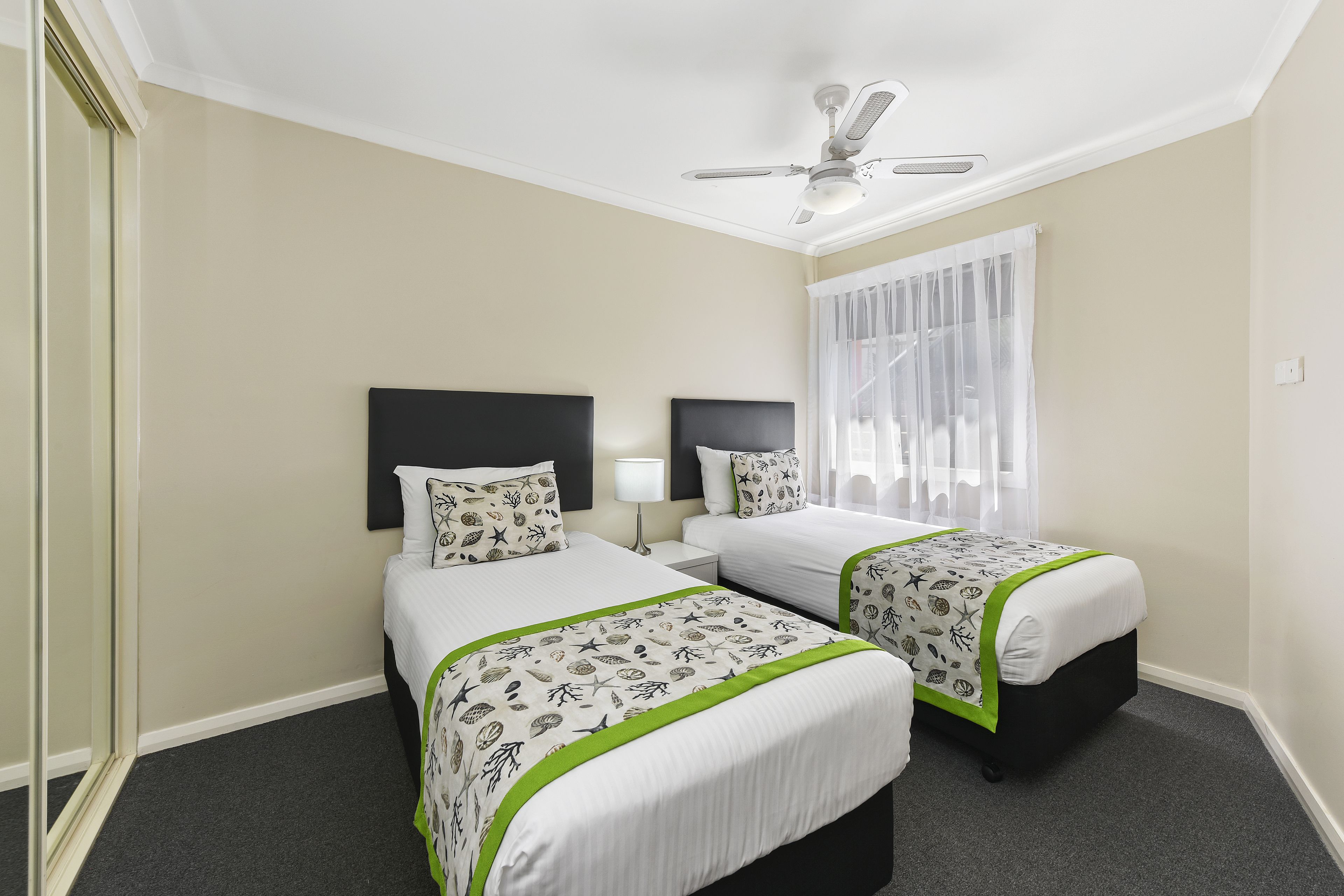 A hotel room with two beds, a ceiling fan, a window with sheer curtains, and a carpeted floor.