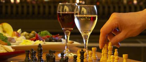 Un tablero de ajedrez con piezas, una copa de vino tinto y una mano moviendo una ficha.