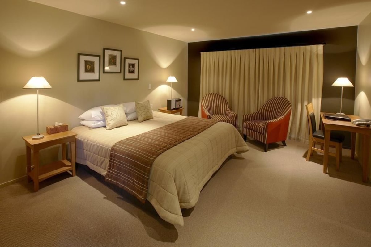 A hotel room with a bed, two bedside tables, a chair, a desk, and a large window with curtains.