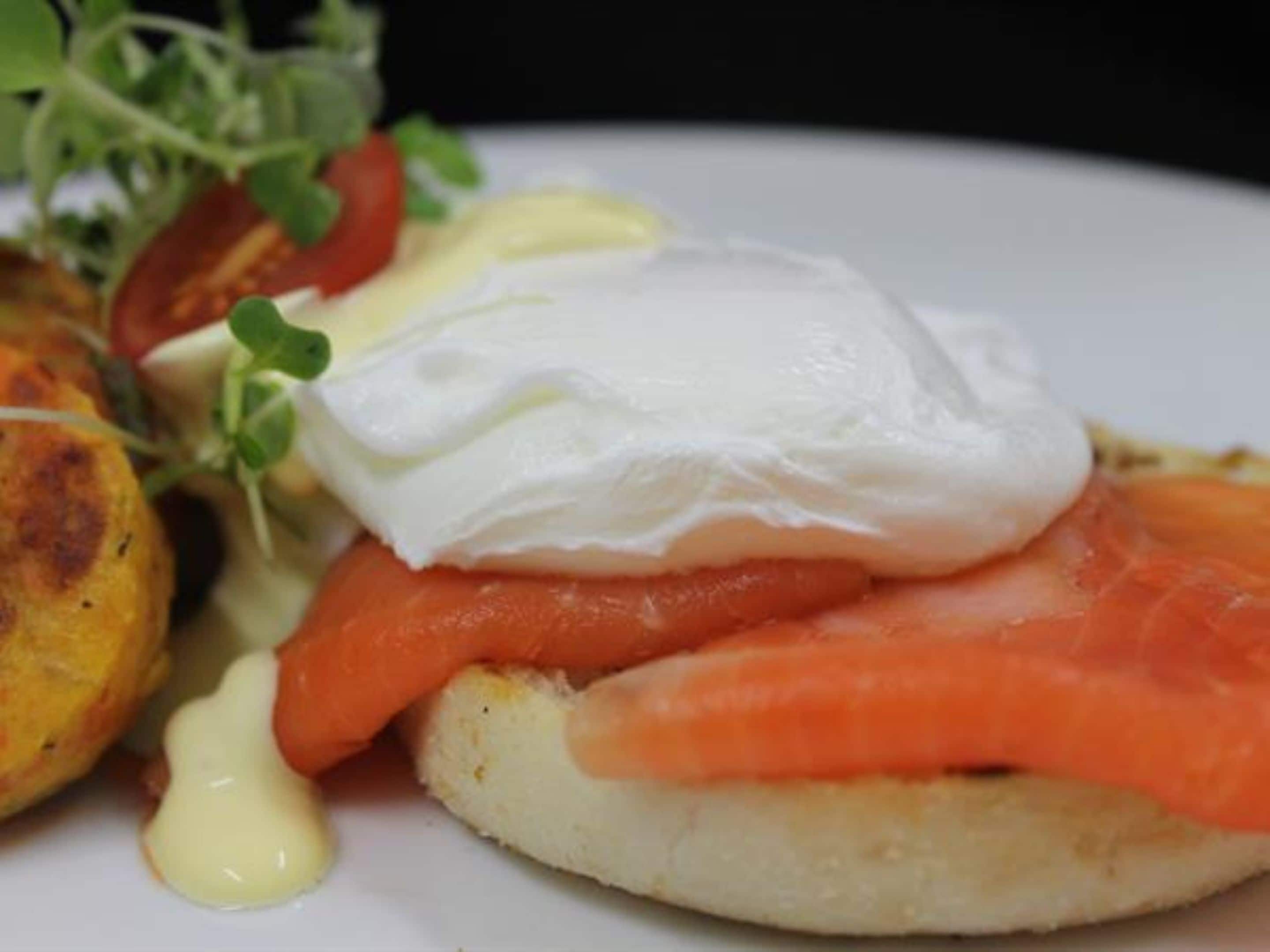 Un plato con un huevo escalfado, salmón ahumado y un huevo escalfado sobre tostada, acompañado de vegetales verdes y salsa.