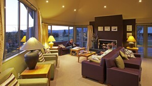 A spacious living room with a fireplace, comfortable seating, and large windows offering a scenic view. - Te Weheka Hotel Fox Glacier  (Fox Glacier)