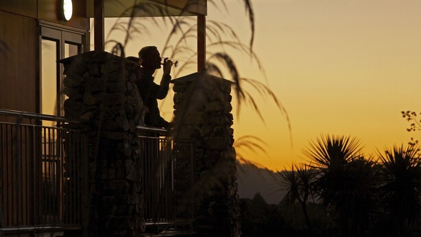 A person using binoculars on a balcony with stone pillars, overlooking palm trees and a sunset. - Te Weheka Hotel Fox Glacier  (Fox Glacier)