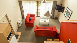 A compact hotel room with a red sofa, a small table, a TV mounted on the wall, and a view through the door.
