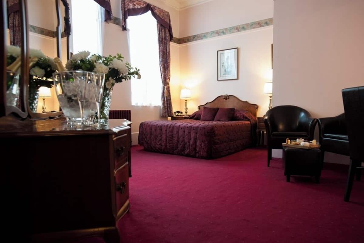 A hotel room with a bed, a dresser, a chair, a lamp, and a vase with flowers.