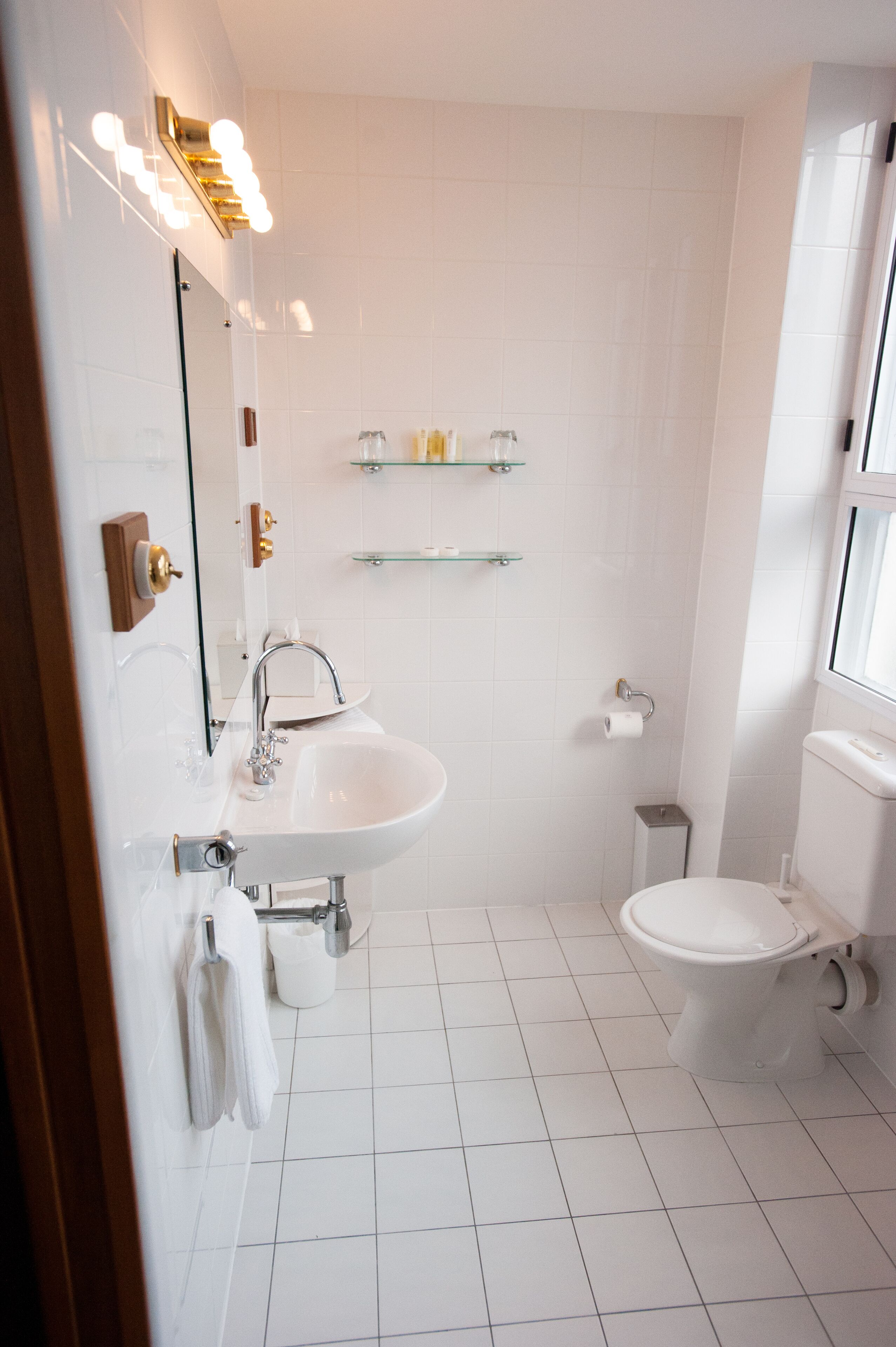 A bathroom with a sink, mirror, and toilet. There are shelves above the sink and a window to the right.